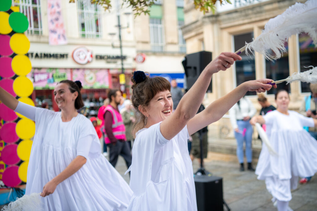 Women in white flowing dresses dance in a town centre with feathers, smiling