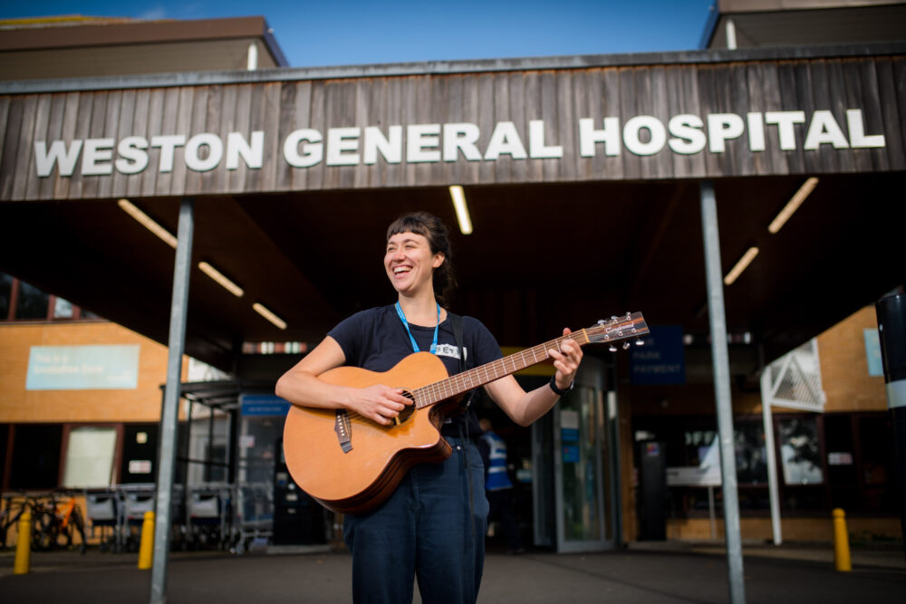 Woman playing a guitar and smiling in front of Weston General Hospital.