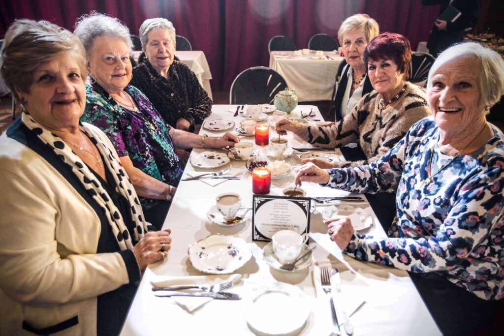 6 older women looking to the camera. They are sitting round a dinner table, waiting to be served. Candles are lit. They look happy
