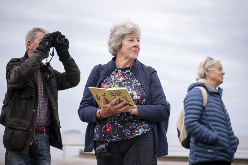 Woman holds a bird book on the beach, looking to her left. On her right is a man looking at the sky through binoculars. There is another women beside her on her left