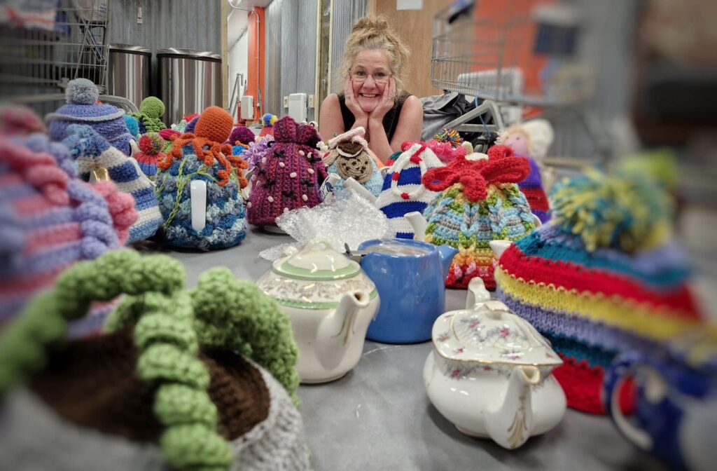 A woman sits at a table, smiling, cupping her chin in her hands. She is surrounded by colourful tea cosies and tea posts