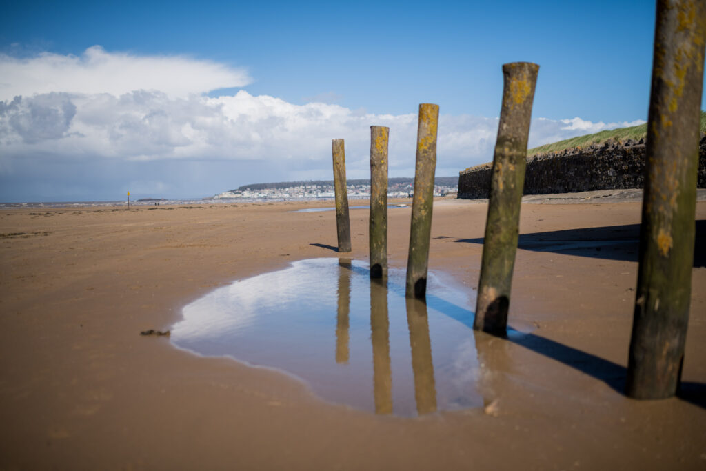 Weston-super-Mare beach. 3 posts, in the sand with a headland behind. Sea water gathers by the posts, the blue sky and a few clouds reflecting