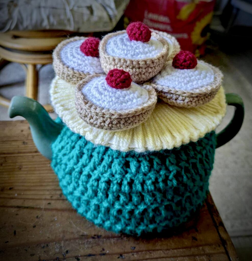 A teapot in a cosy. The knitted is knitted with woollen bakewell tarts on a plate on top