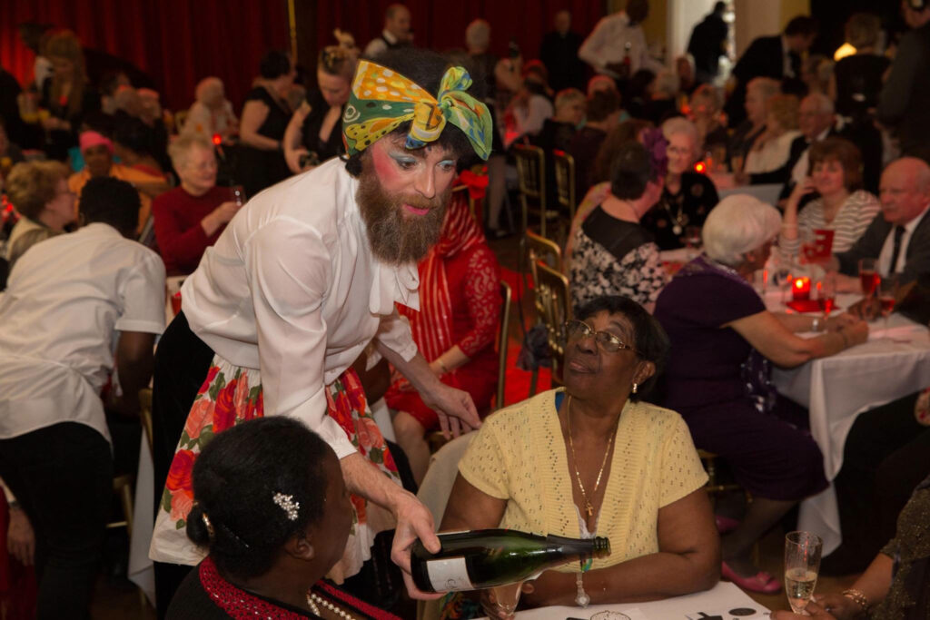 Man with beard and colourful headscarf, with make up on, serving wine to two women at a table in a room full of people, also at tables