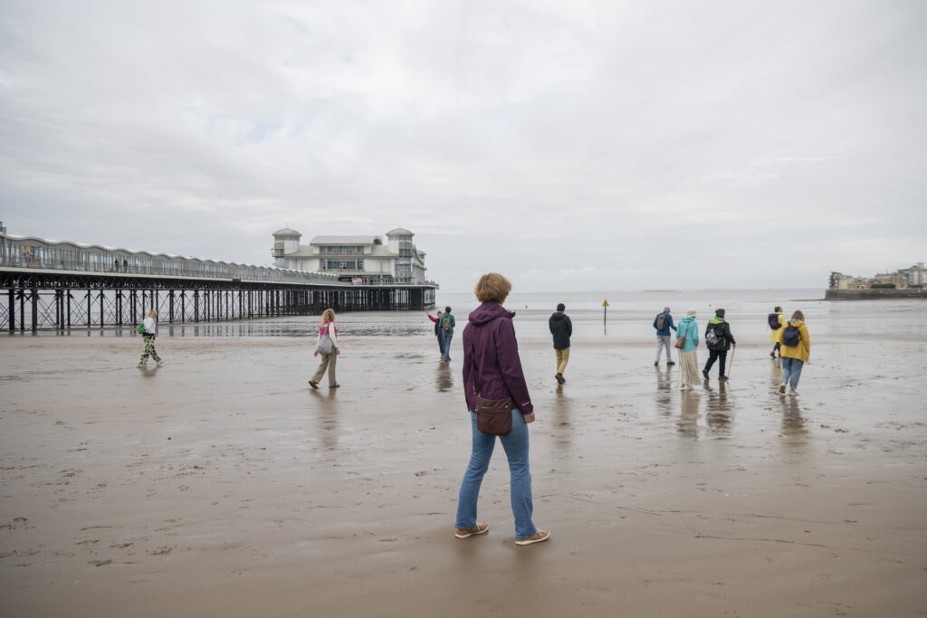 People standing on the beach, some walking. The Grand Pier in Weston-super-Mare is behind them