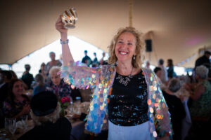 Woman smiling holding up a glass. She is wearing a sequin top in a large, open tent with people all around her