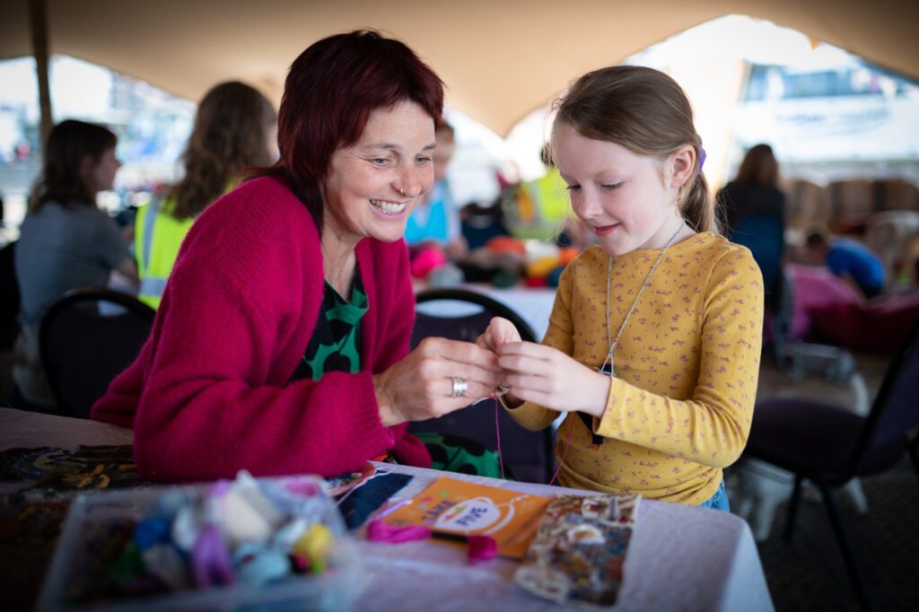 A woman is smiling as she helps a young girl with crafts. They are in a large open tent