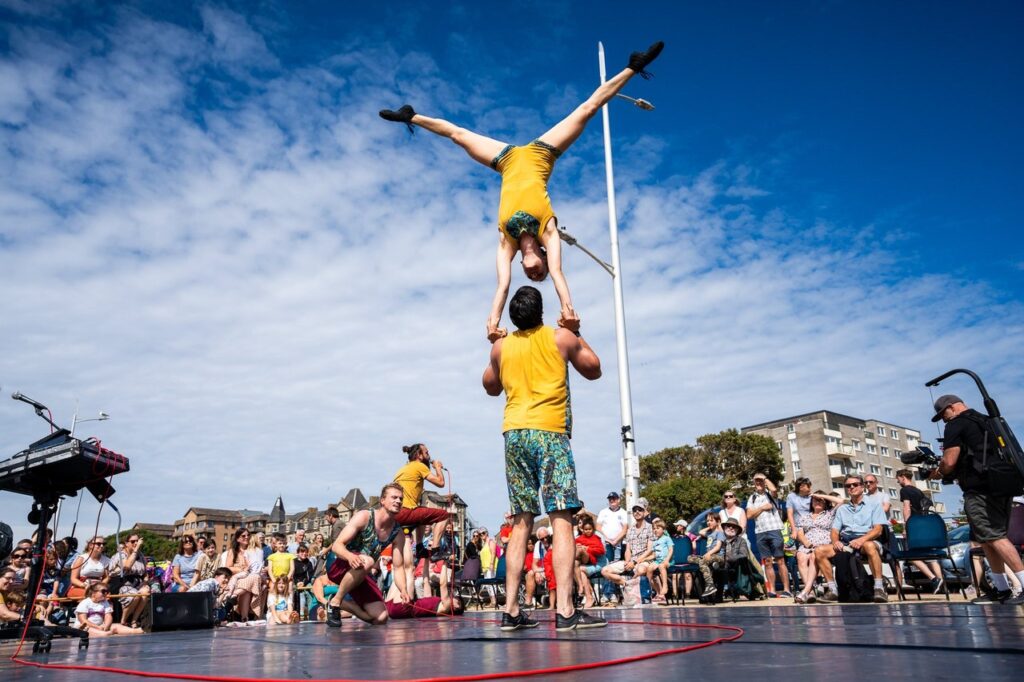 A circus troupe perform on stage on a sunny day. One man signs to the audience, while another looks at a couple on stage, who are doing a trick - a man is holding a woman upside down, above him