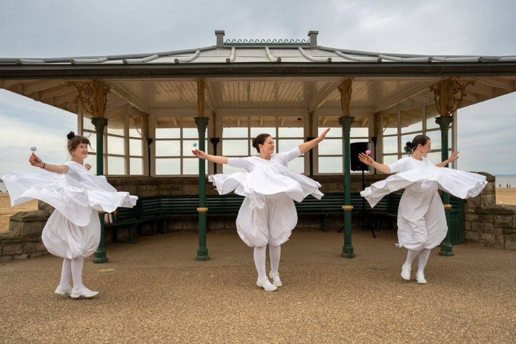 Three women, dressed in white, arms outstretched, with pretend microphones in their hands. Their clothes are spinning around them as they stand in front of a Victorian band stand on the seafront