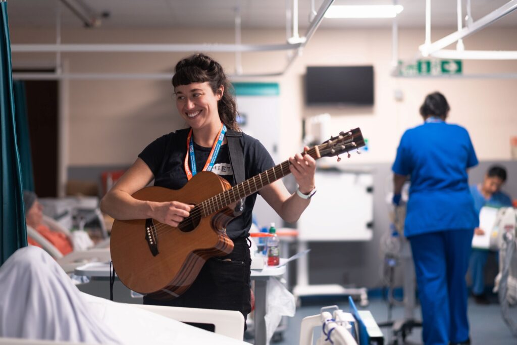 Woman in a hospital ward standing by a bed, smiling and and strumming a guitar