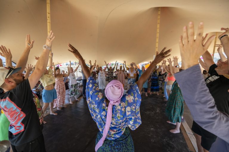 A crowd of people in a circle raise hands in celebration and dance. They are under a large stretch tent