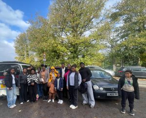 A group of 13 people opf all ages are in a car park under a large tree, they look to camera